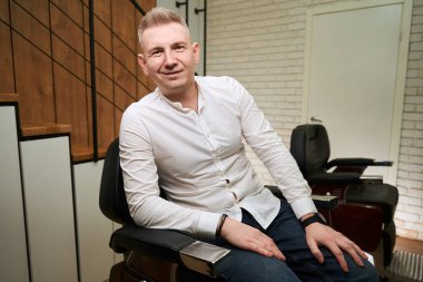 Açık tişörtlü, gülümseyen sarışın profesyonel bir koltukta oturuyor, içeride mükemmel ışıklandırma var.