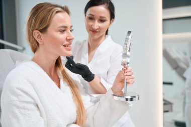Bir kozmetoloji kliniğinin hastası yaşlanmayı önleme prosedürlerinden sonra yüzünü aynada muayene eder.