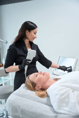 Uzman estetisyen, cihazı gençleştirici bir prosedür için ayarlar, müşteri bornozla yatar.