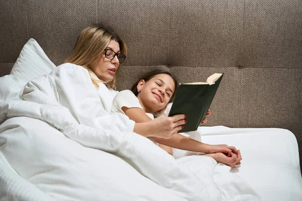戴眼镜的女人躺在床上 微笑着的女儿 看着卧室里的书本 — 图库照片