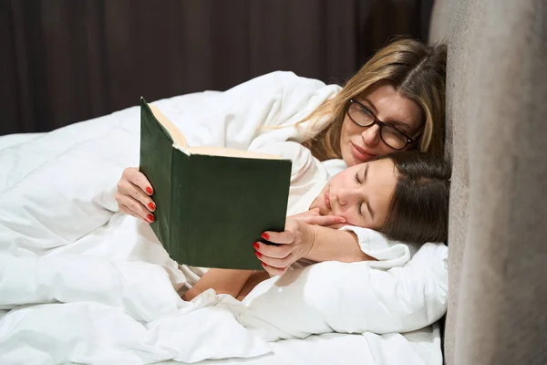 Madre Gafas Tumbada Cerca Chica Dormida Mirando Libro Dormitorio —  Fotos de Stock