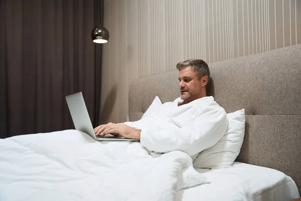 Hombre Adulto Con Abrigo Blanco Relajándose Cama Escribiendo Portátil Motel —  Fotos de Stock