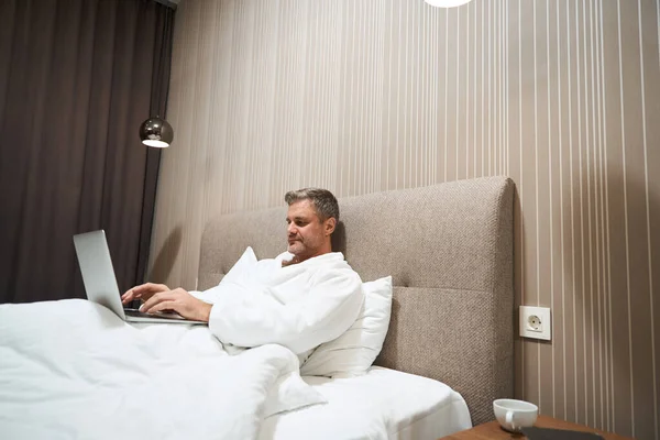 Hombre Adulto Con Abrigo Blanco Descansando Cama Escribiendo Portátil Hotel —  Fotos de Stock