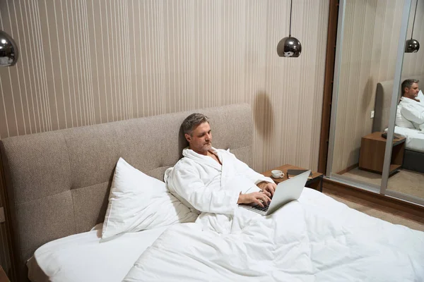 身穿白色外套的男性躺在床上 在汽车旅馆的笔记本电脑上打字 — 图库照片