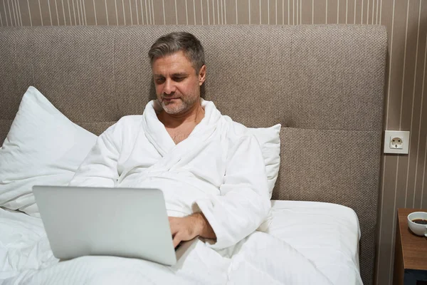 Dorosły Mężczyzna Białym Płaszczu Relaksuje Się Pracuje Laptopie Pokoju Hotelowym — Zdjęcie stockowe