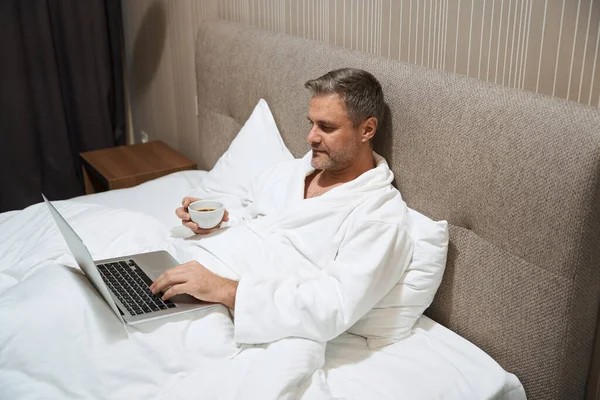 Bărbat Haină Albă Întins Pat Tastând Calculator Ținând Ceașcă Motel — Fotografie, imagine de stoc