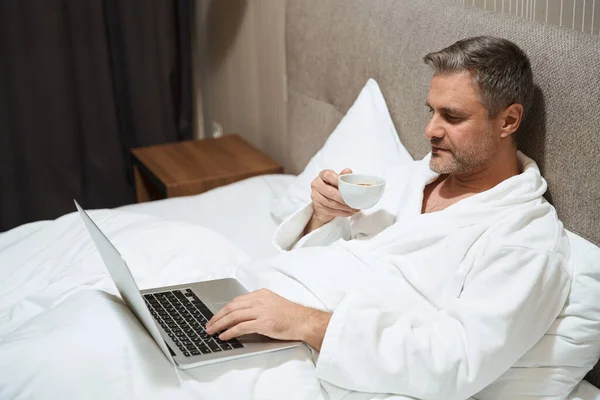 Взрослый Мужчина Белом Халате Отдыхает Кровати Печатает Ноутбуке Пьет Кофе — стоковое фото