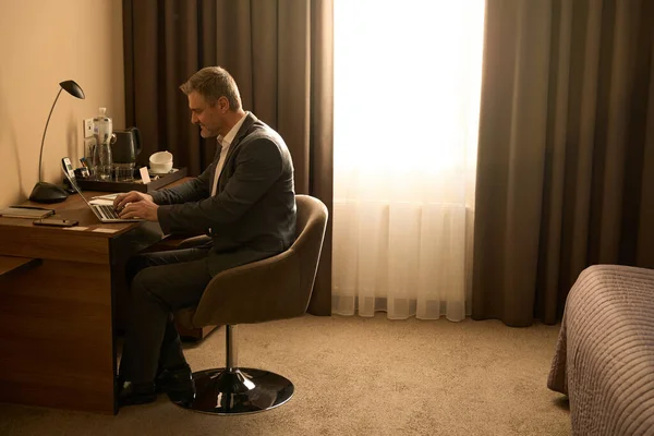 穿着西装的成年男性坐在酒店房间的椅子上 在电脑上打字 — 图库照片