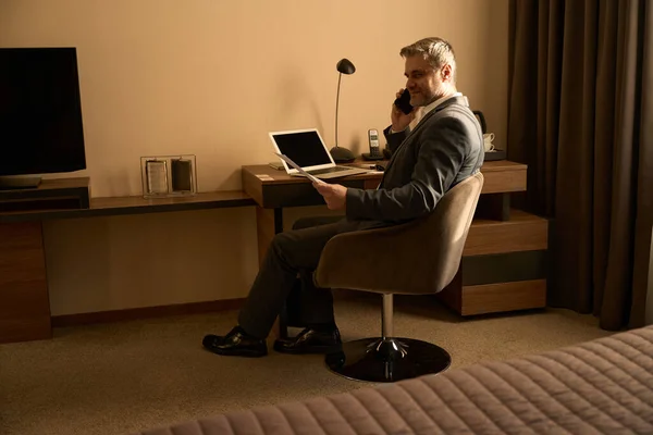 大人のビジネススーツの男が椅子に座って電話で話し ホテルの部屋で書類を持っています — ストック写真