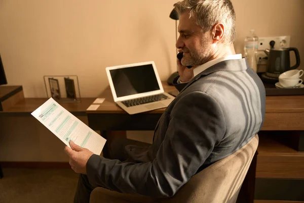 身着西装的男性坐在笔记本电脑前 打电话 在汽车旅馆里拿着文件 — 图库照片
