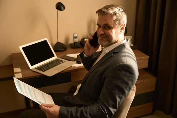 身着西装的成年人坐在笔记本电脑旁 用智能手机打电话 在酒店房间里拿着文件 — 图库照片
