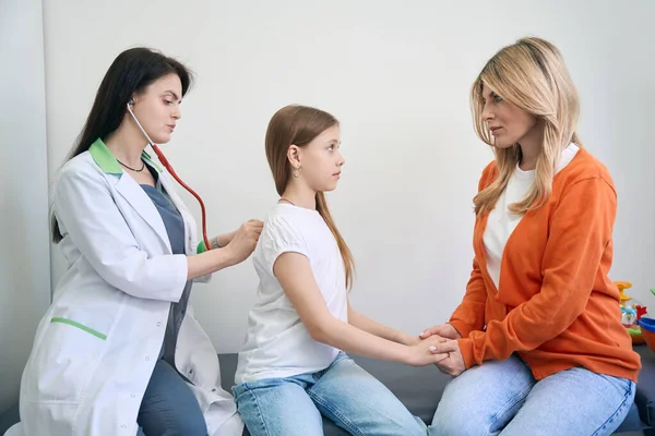 严重的母亲牵着不健康的小女孩的手 而合格的儿科医生则用听诊器和体格检查检查她的肺 — 图库照片