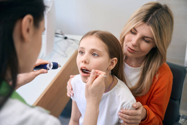 Woman laryngologist examining little girl throat using sepcial stick and pocket light, health check-up