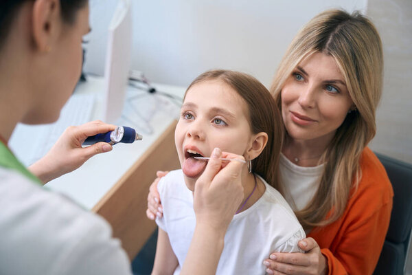 Female ear, nose and throat specialist examining throat of little girl sitting on mothers knees with complains of sore throat, health check-up