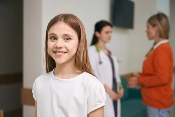 彼女の健康診断の結果を医師と話し合いながら カメラと笑顔を見て小児科の幸せな女の子の患者 — ストック写真