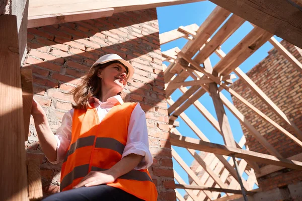 安全ベストとハードハットで木製のはしごに立っている女性の主建設労働者と建物を見て 請負業者の仕事に満足 — ストック写真