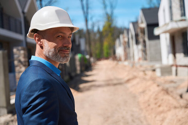 Smiling building engineer in hardhat walking along construction site, checking building stage, planning landscape design, successful housing development