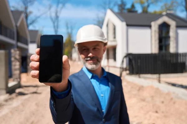 硬い帽子をかぶってスマートフォンのディスプレイを見せながら笑顔のフォアマン 建設現場に立って準備ができてタウンハウスの3Dモデルを示します — ストック写真