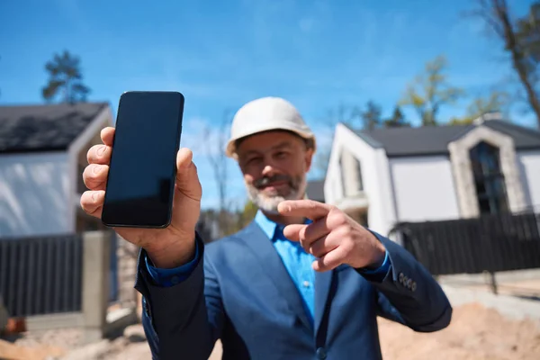 建設現場に立つスマートフォンのディスプレイで指を指すハード帽子の男の建物の管理者を笑顔 住宅の販売とレンタル — ストック写真