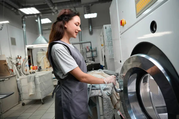 汚れやリフト汚れを除去するために 化学溶剤を使用した乾燥洗浄機に服を入れて笑顔の女性ランドリーサービス事業者 — ストック写真