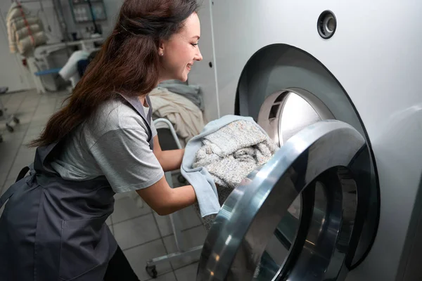 Woman laundry service worker loading big professional washing machine with clothes, delicate fabrics dry cleaning