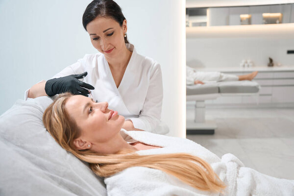 Молодой врач в форме выполняет процедуру инъекции вокруг глаз клиента, эстетик использует тонкую иглу