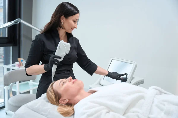 Specialist Cosmetologist Adjusts Device Rejuvenating Procedure Client Lies Cozy Bathrobe — Stock Photo, Image