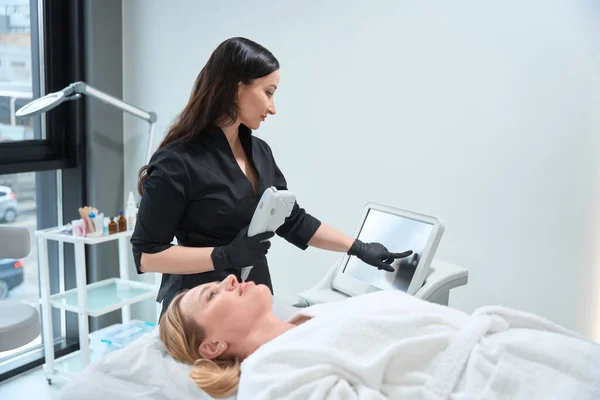 Woman Adjusts Device Rejuvenating Procedure Client Lies Bathrobe — Stock Photo, Image