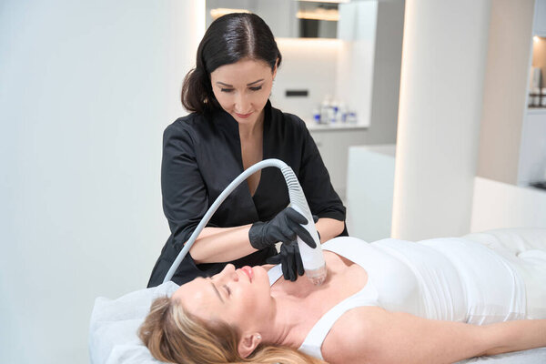 Блондинка лежит на косметологическом диване во время процедуры подъема РФ в зоне декольте, эстетик использует современное оборудование