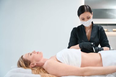 Siyah üniformalı bir kadın müşterinin karnına iğne yapar. Bir estetik uzmanı koruyucu eldivenler ve maske giyer.