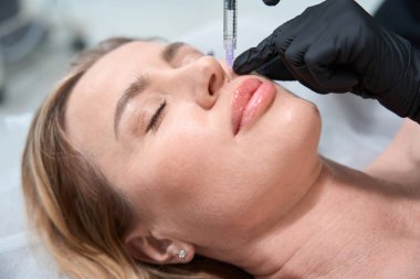 Kozmetik uzmanı sarışın bir müşteriye güzellik iğneleri yapıyor. Bir kadın kozmetoloji kanepesinde yatıyor.