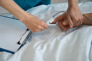 Doktorun, klinikte yatakta yatan erkek hasta parmağına nabız oksimetresi uyguladığı yakın plan.