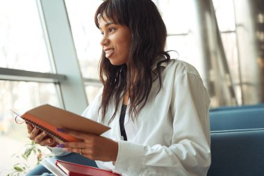 Pencereden bakan ve modern odada elinde fotokopi kitabı tutan mutlu kadın.