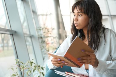Gündelik giysiler içinde güzel bir kadın pencereden dışarı bakıyor ve modern odada fotokopi kitabını tutuyor.