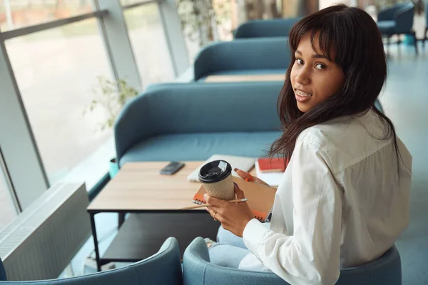 微笑的女人拿着咖啡 坐在舒适的室内椅子上 转身对着相机 — 图库照片