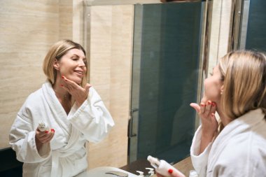 Beyaz önlüklü gülümseyen kadın aynanın yanında duruyor ve yansımaya bakıyor, tuvalette tenine dokunuyor.