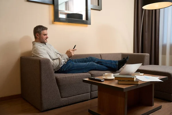 快適なソファでリラックスしながら 携帯電話を閲覧しながら ホテルの部屋で男性の通過時間のサイドビュー — ストック写真