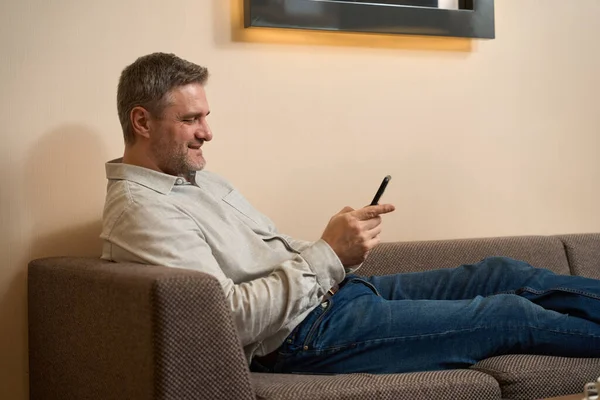 微笑的男人在舒适的沙发上放松和看手机的侧视图 — 图库照片
