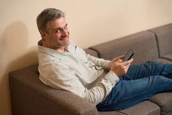 男人一边在舒适的沙发上一边浏览手机一边看相机的侧视图 — 图库照片