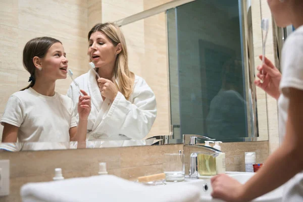 身穿白衣的母亲和站在镜子旁边拿着牙刷的女孩在宾馆里互相望着 — 图库照片
