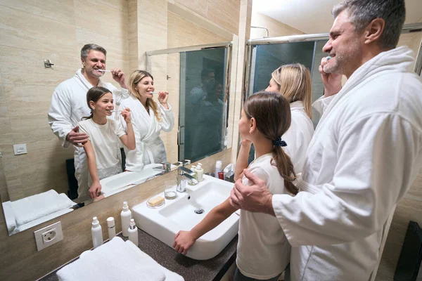 微笑着的男士 漂亮的女士和穿着白色浴衣的少女 一边刷牙 一边照着镜子 — 图库照片