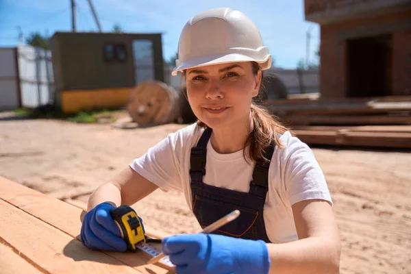 笑顔の女性建設労働者は 測定テープで木製の梁の対策を取ります 材料をソートし 屋根フレームの建設のための最高のビームを選択します — ストック写真