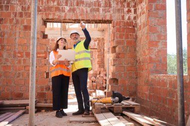 Çatı kirişleri dizaynını planlı dizüstü bilgisayarla tartışan inşaat mühendisleri inşaat sahasında yürüyorlar.