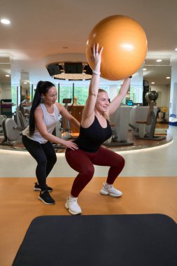 Sarışın bir fitness eğitmeninin gözetiminde egzersiz yapıyor, fitball kullanıyor.