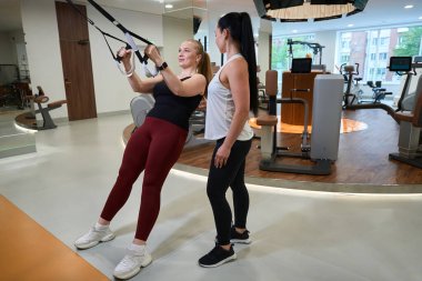 Güçlü genç bir kadın modern bir simülatörde çalışıyor. Deneyimli bir fitness eğitmeni yakınlarda.
