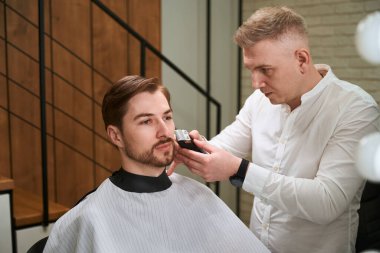 Profesyonel kuaför, kuaför pelerinli müşterisine içeride berber makinesi kullanarak hizmet veriyor.