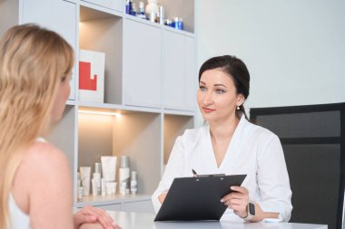 Kadın kozmetikçi, kozmetoloji kliniğindeki bir müşterisiyle bakım ürünleri raflarında iletişim kuruyor.