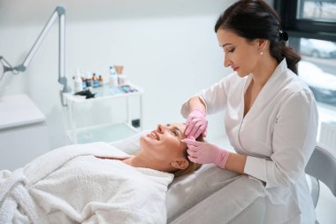 Kozmetik uzmanı hastaların çalışma alanlarını işaretlemek için özel bir işaret kullanıyor. Kadın ise kozmetoloji kanepesinde.