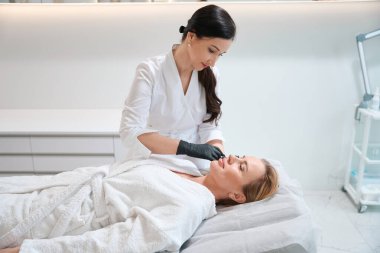 Estetisyen güzel sarışın bir kadına güzellik iğneleri yapıyor. Bornozlu bir kadın kozmetoloji kanepesinde yatıyor.