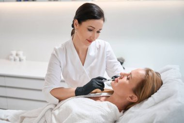 Bir kozmetoloji kliniğinin çalışanı hastaya güzellik iğnesi yapar, bir kadın koruyucu eldivenlerle çalışır.